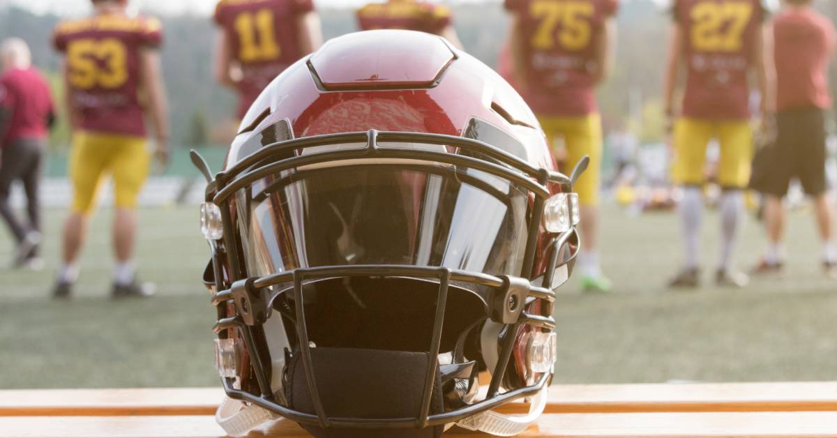 A burgundy and black football helmet resting on a sideline bench. The team is standing together in the background.